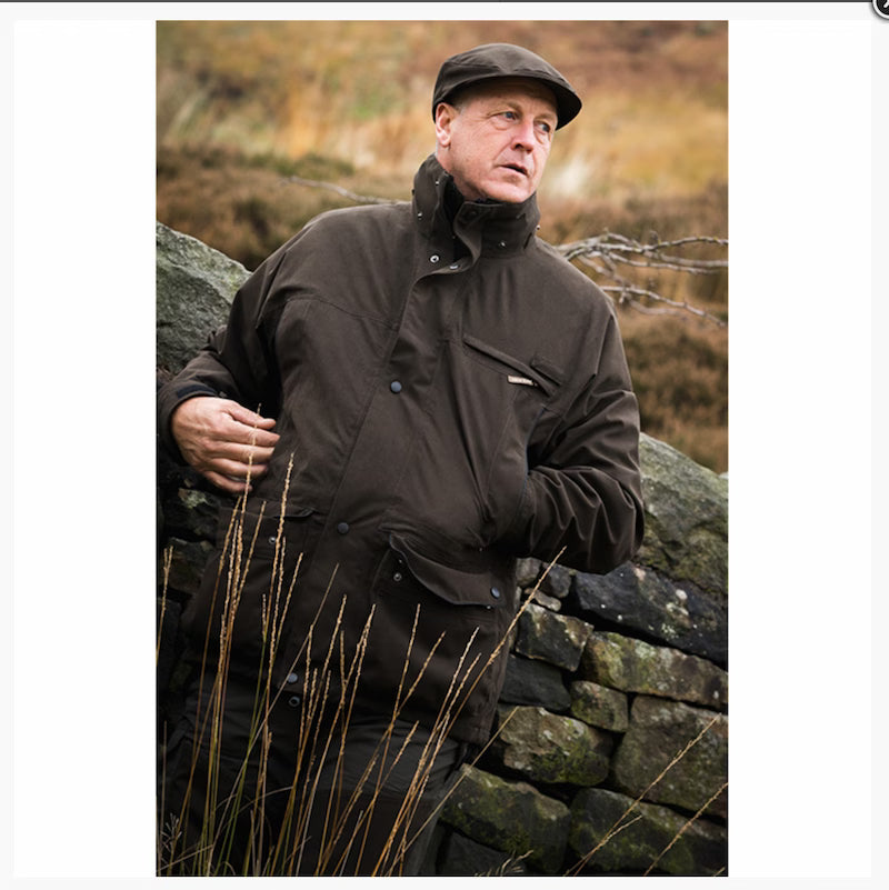 Man in a dark jacket and cap standing by a stone wall with a natural background