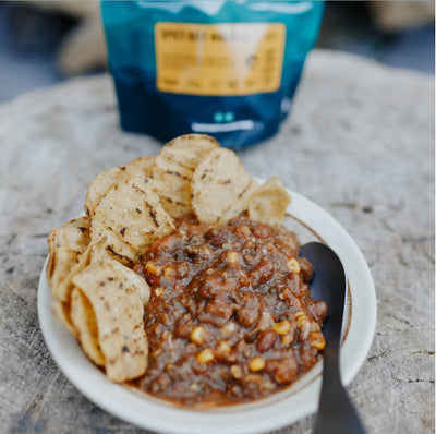 Bowl of chili with tortilla chips on a stone surface, with a blue bag in the background.