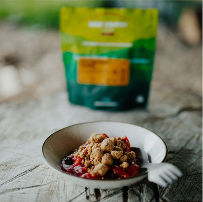 Small bowl of dessert with crumble topping on a wooden surface, with a green bag in the background.