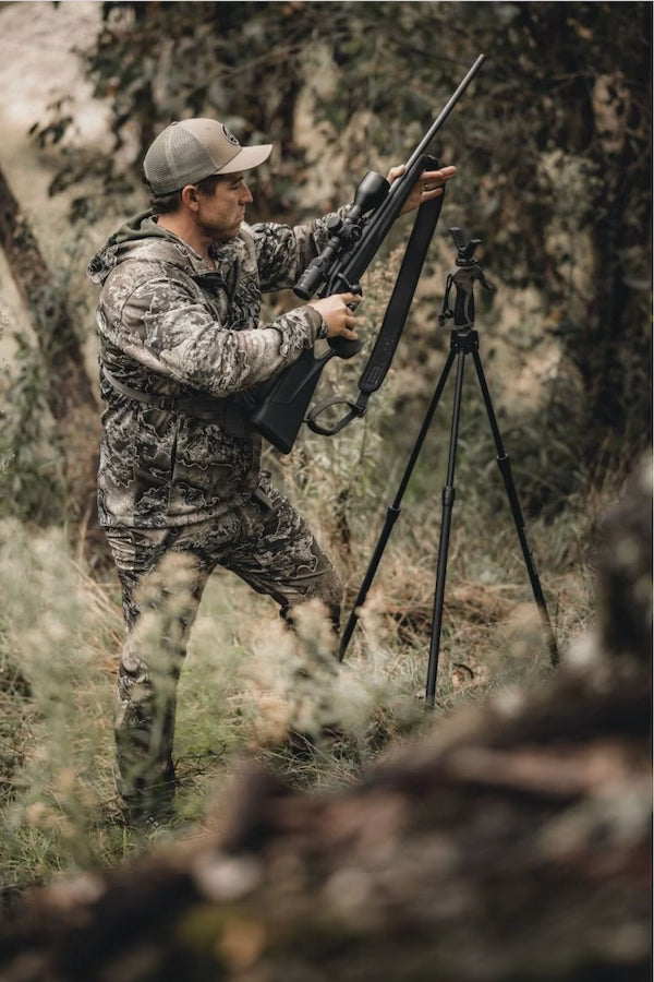 Hunter in camouflage gear with a rifle and tripod in a forest setting