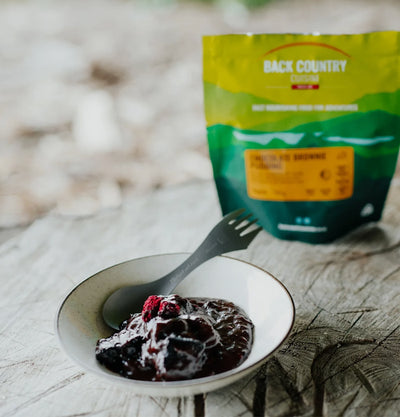 Pudding in a bowl with a fork and a 'Back Country' package in the background on a wooden surface.