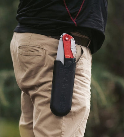 Person wearing a black leather sheath with a red pocket knife on beige pants against a blurred natural background