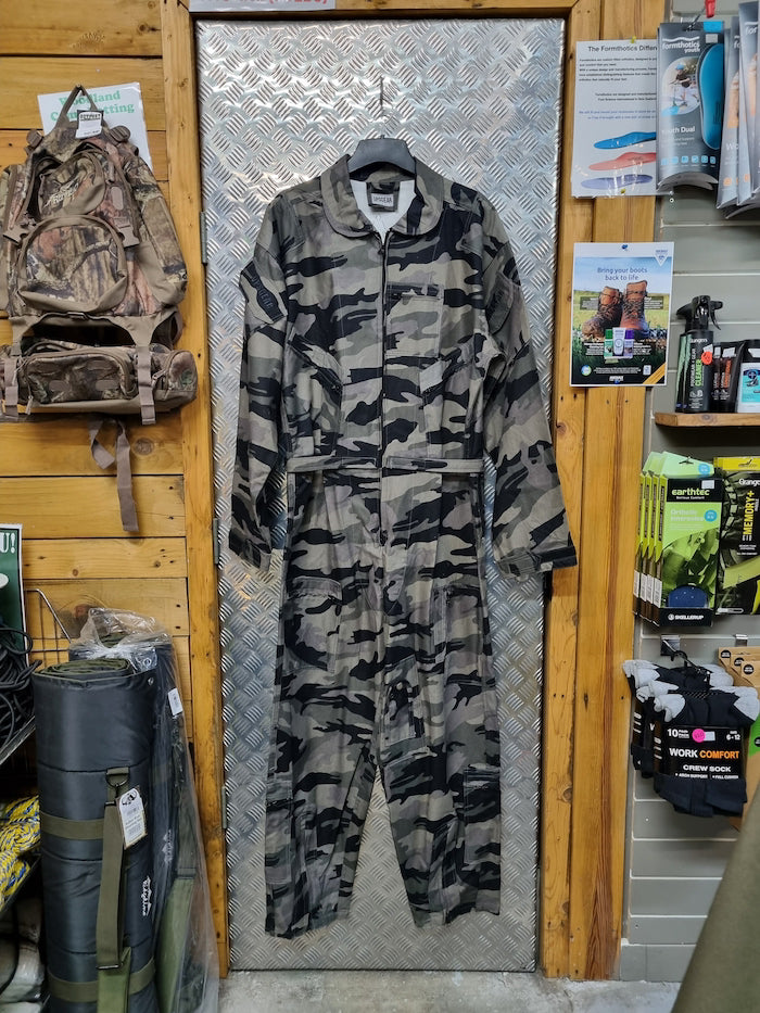 Camouflage jumpsuit displayed on a hanger against a wooden wall with outdoor gear.