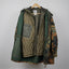 A mixed-material quilted jacket hanging on a wooden hanger against a diamond plate metal wall. The jacket features a tan quilted front panel, dark green fabric on the sleeves and side pockets, and a camouflage pattern on the right sleeve.