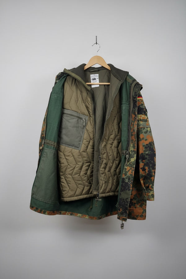A mixed-material quilted jacket hanging on a wooden hanger against a diamond plate metal wall. The jacket features a tan quilted front panel, dark green fabric on the sleeves and side pockets, and a camouflage pattern on the right sleeve.