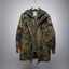 A hooded camouflage jacket hanging on a wooden hanger against a gray gradient background. The jacket features a German Flecktarn-style digital camouflage pattern in green, brown, and black, with an olive green inner lining and a dark green hood.
