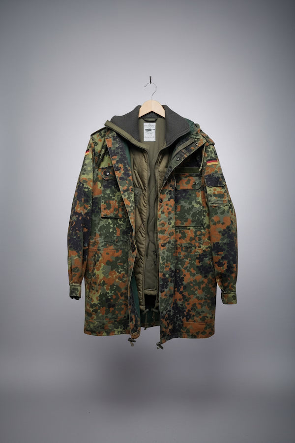 A hooded camouflage jacket hanging on a wooden hanger against a gray gradient background. The jacket features a German Flecktarn-style digital camouflage pattern in green, brown, and black, with an olive green inner lining and a dark green hood.