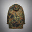 A hooded camouflage jacket shown from the back, hanging against a gray gradient studio background. The pattern mixes olive green, brown, black, and tan in a digital camouflage print.
