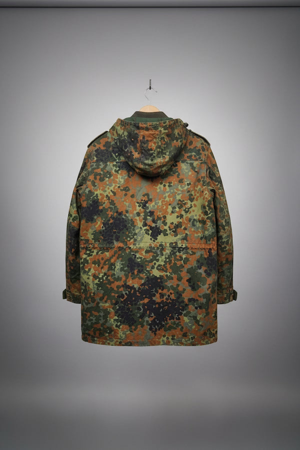 A hooded camouflage jacket shown from the back, hanging against a gray gradient studio background. The pattern mixes olive green, brown, black, and tan in a digital camouflage print.