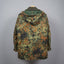 A hooded camouflage jacket shown from the back, hanging on a wooden hanger against a plain gray studio background. The camouflage pattern mixes olive green, brown, black, and tan tones, with a solid olive green hood lining.