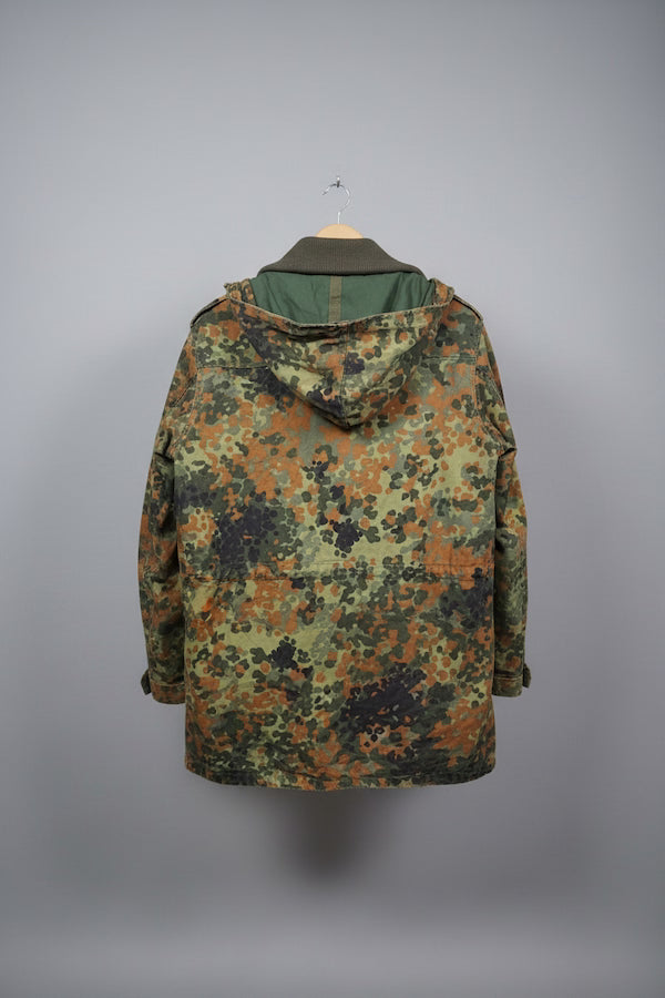 A hooded camouflage jacket shown from the back, hanging on a wooden hanger against a plain gray studio background. The camouflage pattern mixes olive green, brown, black, and tan tones, with a solid olive green hood lining.