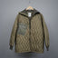 An olive green quilted jacket with a zippered front, a large chest pocket, and a hood, hanging on a wooden hanger against a solid gray background.