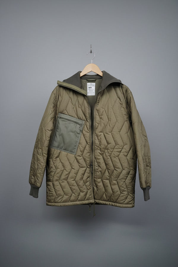 An olive green quilted jacket with a zippered front, a large chest pocket, and a hood, hanging on a wooden hanger against a solid gray background.