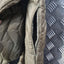 Close-up of a quilted puffer jacket showing a zippered pocket and interior lining in a muted olive-tan color, with a dark diamond-plate surface in the background.