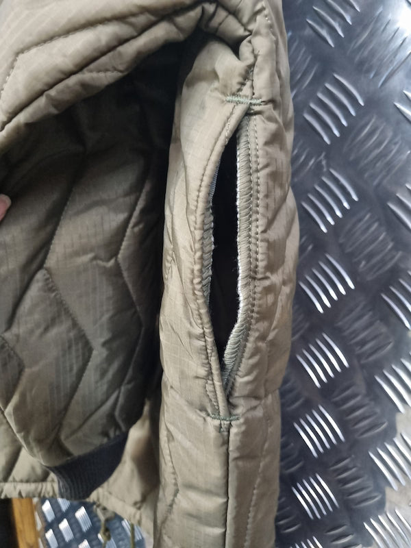Close-up of a quilted puffer jacket showing a zippered pocket and interior lining in a muted olive-tan color, with a dark diamond-plate surface in the background.