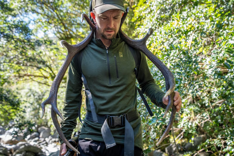 A man in outdoor gear, including a green quarter-zip fleece, cap, and harness, carrying a large pair of antlers over his shoulders in a forest setting.