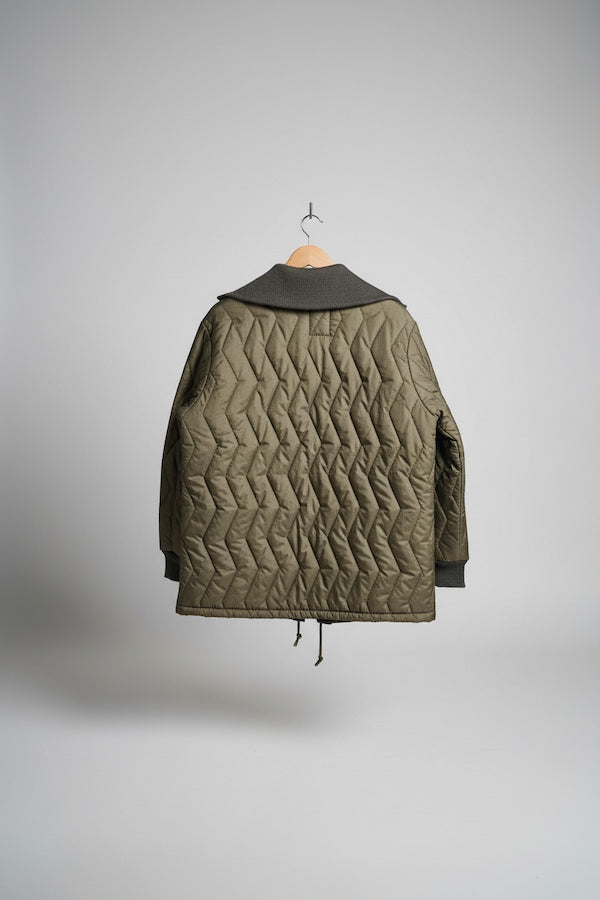 An olive green quilted jacket with a dark gray hood, hanging on a wooden hanger against a diamond plate metal door.