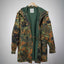 A German Flecktarn camouflage military parka jacket with a green interior lining, hanging on a wooden hanger against a gray gradient background.