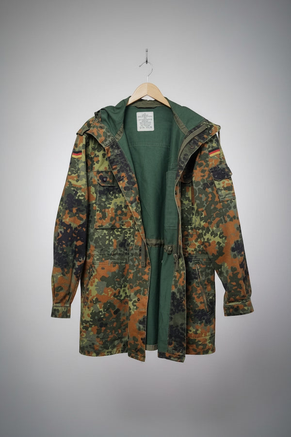 A German Flecktarn camouflage military parka jacket with a green interior lining, hanging on a wooden hanger against a gray gradient background.