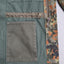Close-up of a vintage camouflage jacket, showing a green canvas body, a brown zipper with a metal pull, and a camouflage lining.