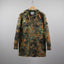A German Flecktarn camouflage military jacket hanging on a wooden hanger against a neutral gray background.