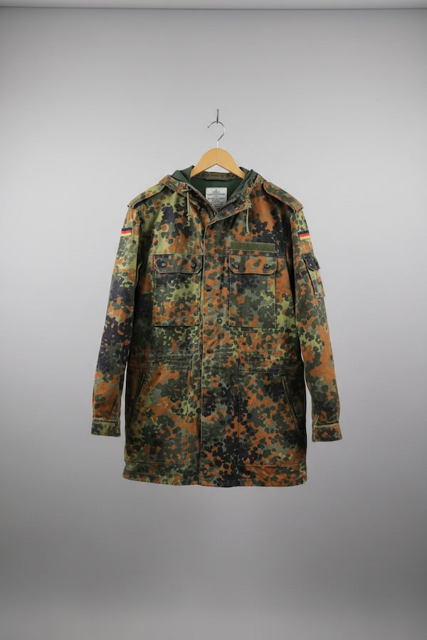 A German Flecktarn camouflage military jacket hanging on a wooden hanger against a neutral gray background.