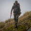 A hiker wearing an olive green hooded jacket, matching pants, and a large backpack with a trekking pole, walking up a mossy grassy hillside in a foggy outdoor setting.