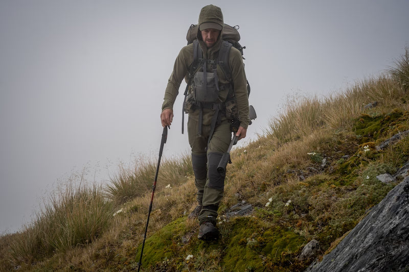 A hiker wearing an olive green hooded jacket, matching pants, and a large backpack with a trekking pole, walking up a mossy grassy hillside in a foggy outdoor setting.