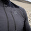 Close-up of a man wearing a dark gray half-zip athletic jacket with a vertical chest zip pocket and a small embroidered logo on the left chest, set against a blurred outdoor beach background.