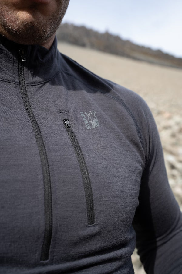 Close-up of a man wearing a dark gray half-zip athletic jacket with a vertical chest zip pocket and a small embroidered logo on the left chest, set against a blurred outdoor beach background.