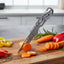 A tactical folding pocket knife with a textured gray handle and silver blade is shown inserted into a wooden cutting board, surrounded by fresh vegetables like sliced carrots, red and yellow bell peppers, and rosemary in a blurred kitchen setting.