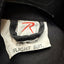 Close-up of a black flight suit interior label with a white tag reading 'SIZE: MEDIUM FLIGHT SUIT' and a red stylized logo, plus partial text 'of New Zealand' at the top.