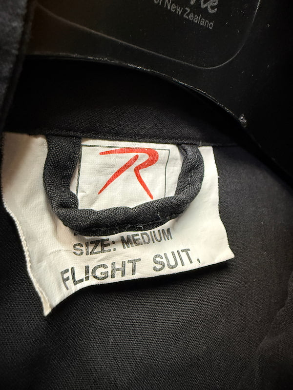 Close-up of a black flight suit interior label with a white tag reading 'SIZE: MEDIUM FLIGHT SUIT' and a red stylized logo, plus partial text 'of New Zealand' at the top.