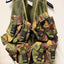 A military-style tactical vest with multiple camouflage pouches and straps, hanging on a black hanger against a plain off-white background.
