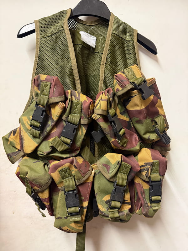 A military-style tactical vest with multiple camouflage pouches and straps, hanging on a black hanger against a plain off-white background.