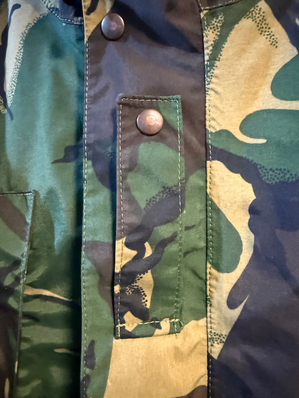 Close-up of a camouflage jacket with green, tan, and dark navy panels, featuring a vertical snap-button placket and a flap pocket with a metal snap.