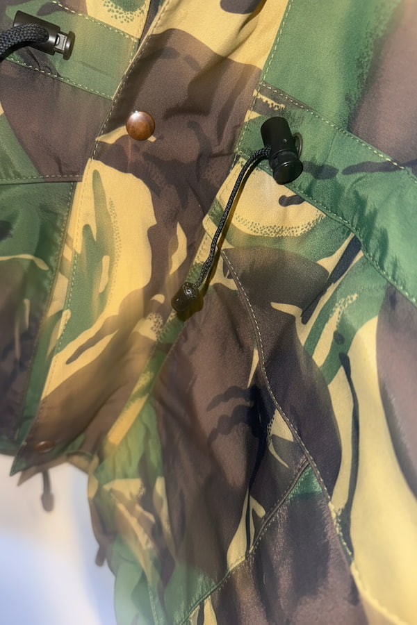 Close-up of a camouflage-patterned garment, likely a jacket or overshirt, featuring green, tan, and dark brown camo print with a snap button and drawstring closure.