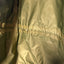 Close-up of a shiny olive green waterproof jacket or rain cover showing the fabric texture, horizontal seam stitching, and elasticized bottom edge.