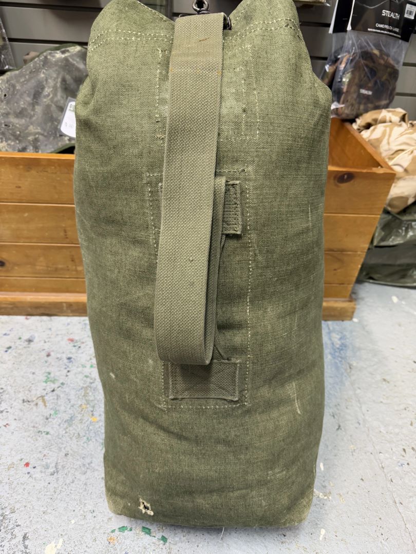 Green military-style duffel bag on a concrete floor with wooden shelves in the background.