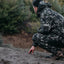 Person in camouflage gear crouching outdoors, possibly hunting or exploring.