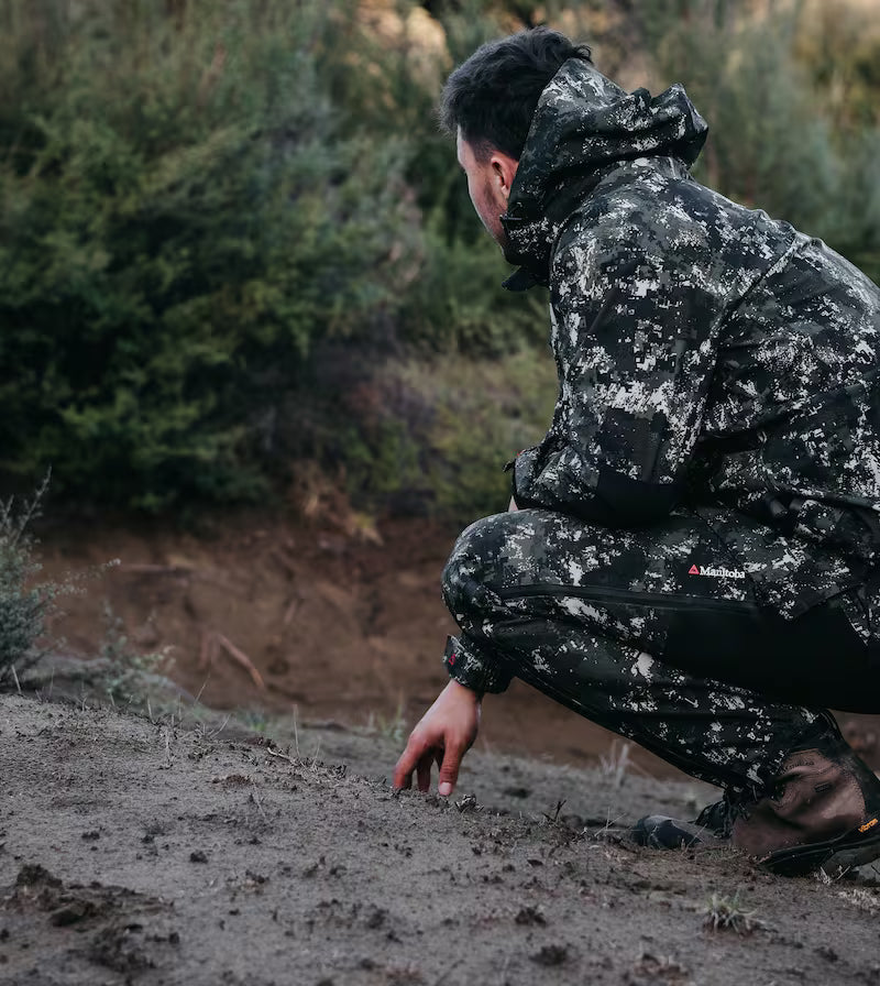 Person in camouflage gear crouching outdoors, possibly hunting or exploring.