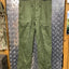 Green military-style pants hanging on a metal surface with camouflage clothing in the background.