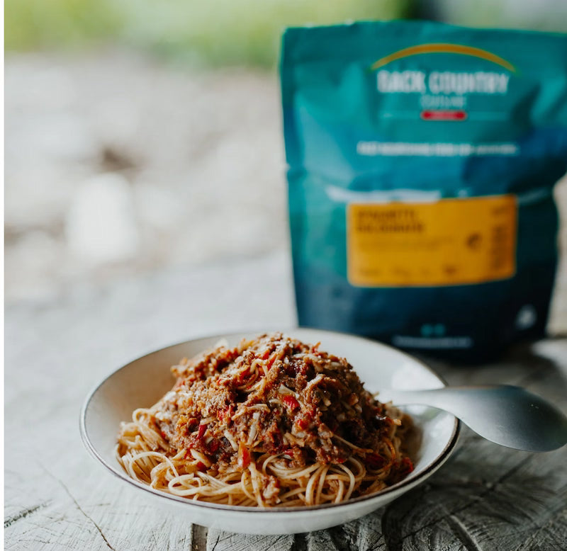 Spaghetti dish with meat sauce on a plate next to a backpack labeled 'Backcountry' on a wooden surface.