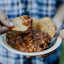 Person holding a bowl of chili with a tortilla chip, blurred blue plaid shirt in the background