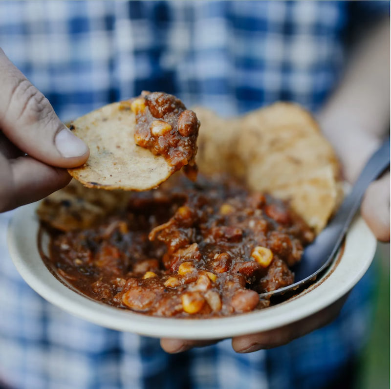 Person holding a bowl of chili with a tortilla chip, blurred blue plaid shirt in the background