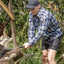 Man in plaid shirt and cap using an axe outdoors with greenery in the background