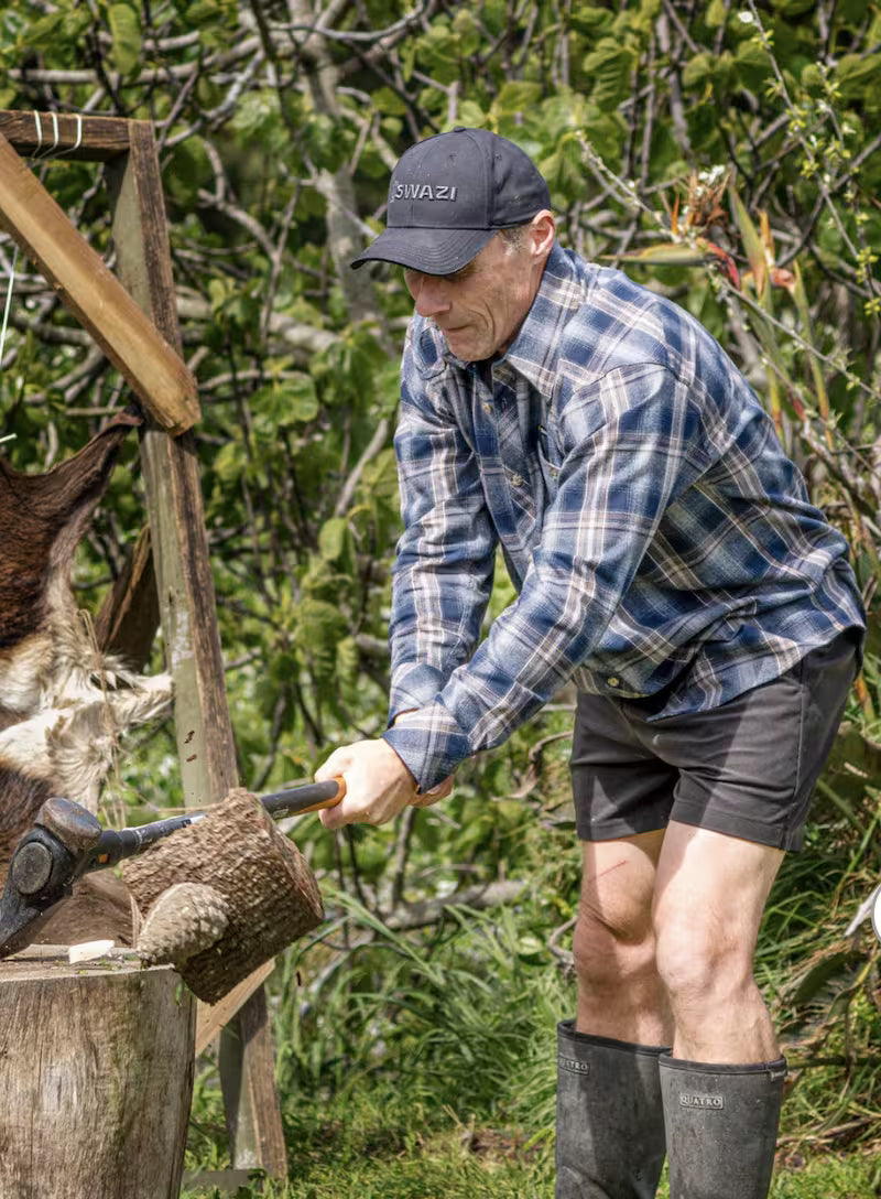 Man in plaid shirt and cap using an axe outdoors with greenery in the background