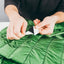 Close-up of a person's hands peeling back the protective paper liner from a black and white adhesive patch, demonstrating how to apply it to a green quilted puffer jacket.