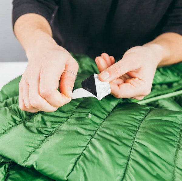 Close-up of a person's hands peeling back the protective paper liner from a black and white adhesive patch, demonstrating how to apply it to a green quilted puffer jacket.