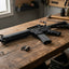 A black tactical rifle laid diagonally across a rustic wooden workbench in a workshop setting, surrounded by tools and hardware.